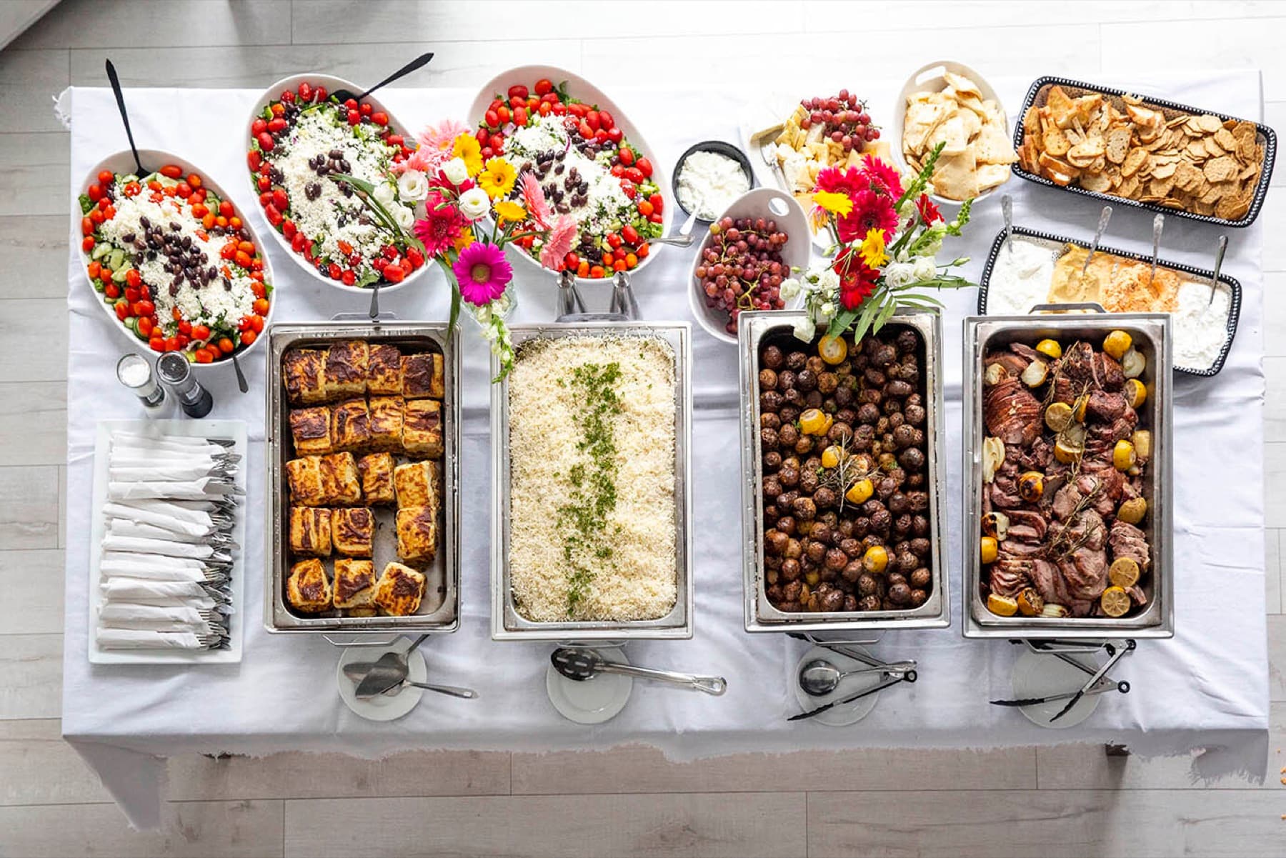 Overhead Greek catering buffet with salads, grilled meats, rice, potatoes, pita, dips, and desserts