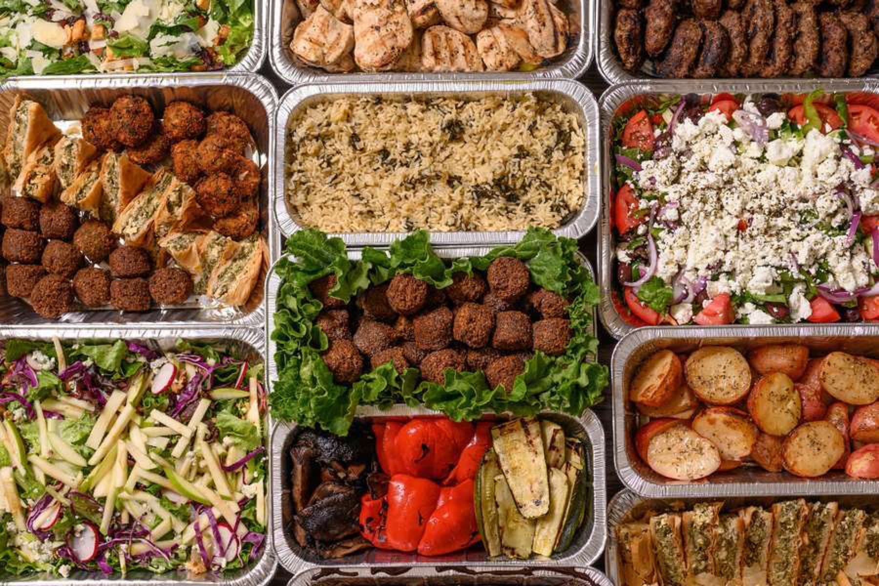 Greek catering package trays with falafel, spanakopita, salads, rice, potatoes, and grilled proteins